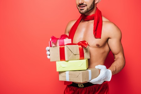 partial view of muscular shirtless sexy man in red scarf holding pile of christmas presents isolated on red backgroundの写真素材