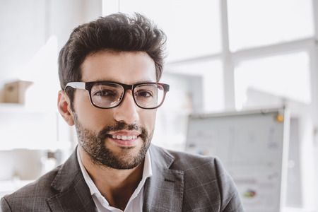 portrait of smiling handsome businessman in glasses looking at camera in officeの写真素材