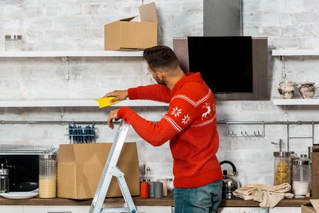rear view of man standing on ladder and wiping dust from shelves during relocation at new homeの写真素材