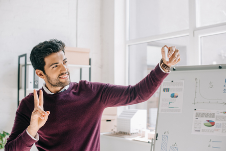 smiling designer in burgundy sweater taking selfie with smartphone and showing two fingers in officeの写真素材