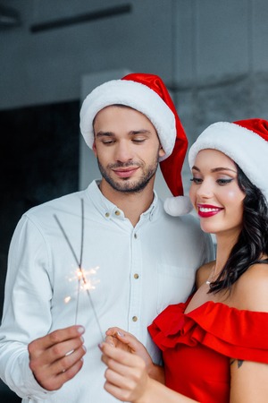 selective focus of young couple in christmas hats celebrating with bengal lights at homeの写真素材