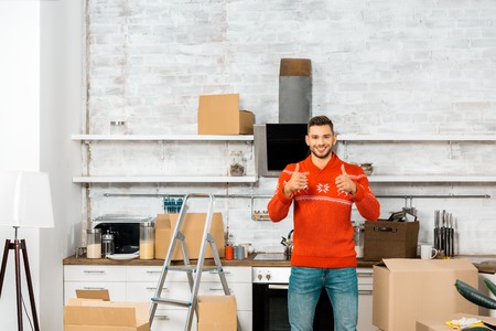 happy young man doing thumbs up in kitchen with cardboard boxes during relocation in new homeの写真素材