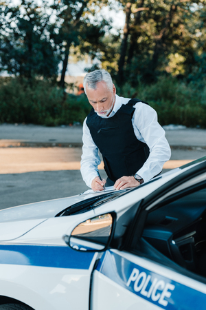 concentrated mature male police officer writing in clipboard on car at streetの写真素材