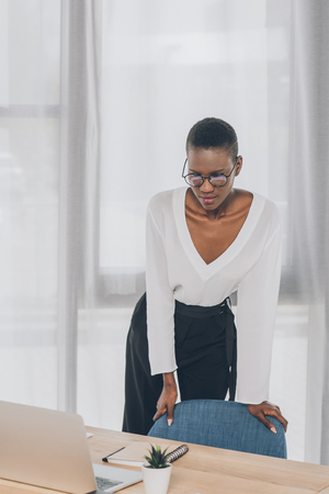 stylish attractive african american businesswoman leaning on armchair and looking at laptop in officeの写真素材