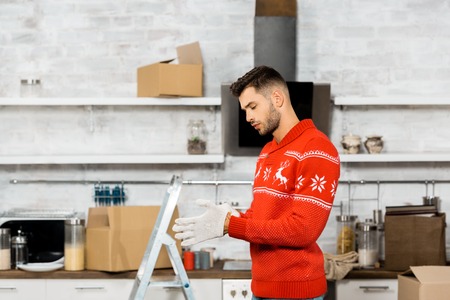 side view of handsome young man putting on working gloves in kitchen with ladder and cardboard boxes...