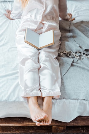 cropped shot of young woman in pajamas sitting on bed with bookの写真素材