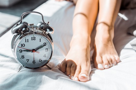 close-up partial view of female feet and alarm clock on bedの写真素材