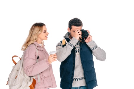 smiling young woman with backpack holding paper cup and looking at handsome man photographing with...