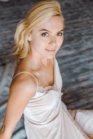 high angle view of beautiful blonde girl sitting and smiling at camera in bedroom