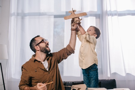 happy father and little son playing with wooden toy plane while modeling together at homeの写真素材