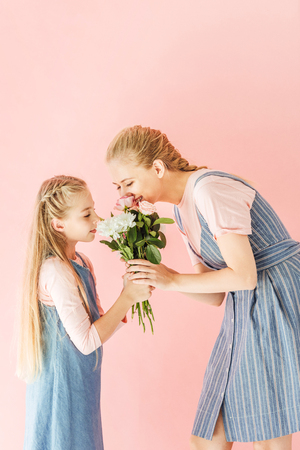 happy young mother and daughter sniffing bouquet together isolated on pinkの写真素材