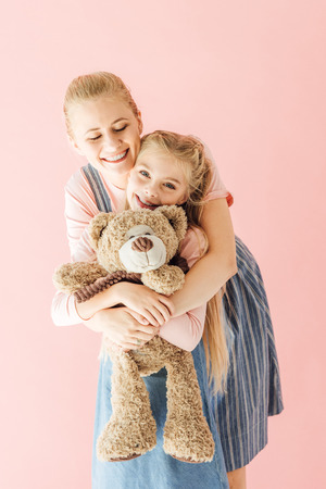 happy mother and daughter with teddy bear embracing isolated on pinkの写真素材