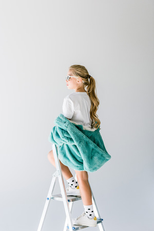 adorable fashionable kid posing in turquoise fur coat on ladder isolated on greyの写真素材