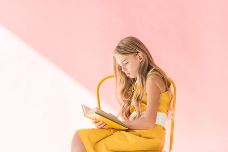 stylish adorable child reading book while sitting on yellow chair on pinkの写真素材