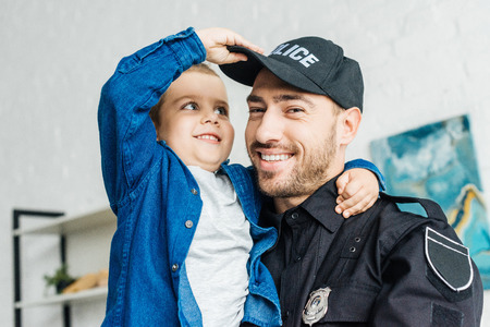 close-up portrait of smiling young father in police uniform carrying his little son and looking at cameraの写真素材
