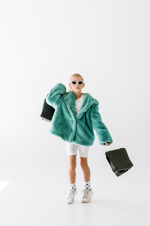 adorable stylish child in turquoise coat and sunglasses jumping with black shopping bags isolated on whiteの写真素材