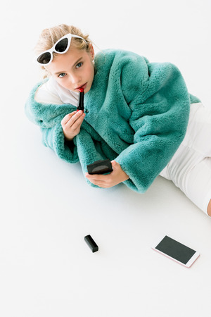 stylish blonde kid in turquoise fur coat applying lipstick with mirror while lying on white with smartphoneの写真素材