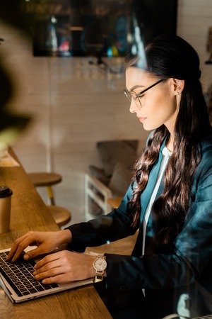 beautiful brunette teleworker in eyeglasses using laptop at restaurantの写真素材