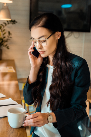 beautiful businesswoman talking on smartphone and holding coffee cup in coffee shopの写真素材