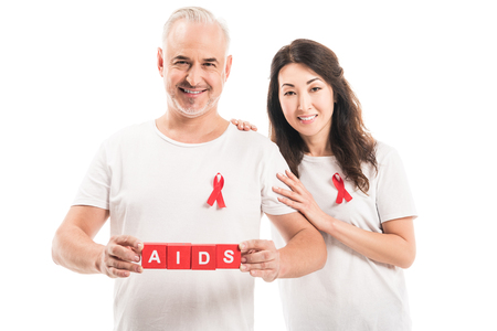smiling adult interracial couple in blank t-shirts with aids awareness red ribbons and blocks with AIDS lettering looking at camera isolated on whiteの写真素材