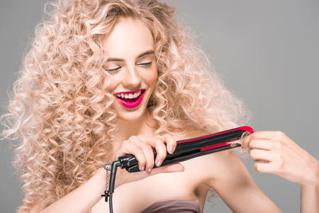 smiling young woman with long curly hair holding hair straightener isolated on greyの写真素材