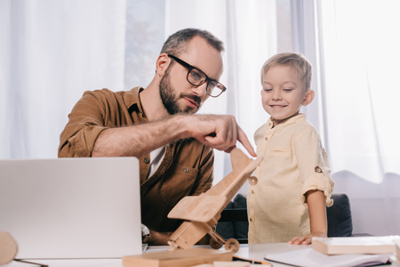 smiling little son looking at father holding wooden plane model at homeの写真素材