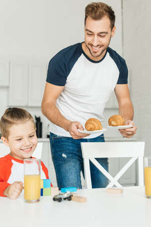 handsome young father serving croissants for breakfast with son while he playing with toysの写真素材