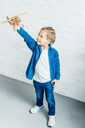 adorable little kid playing with wooden toy airplane in front of white brick wallの写真素材