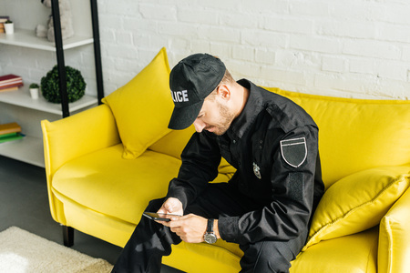handsome young policeman with smartphone sitting on yellow couch