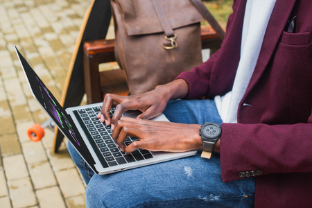 cropped shot of freelancer using laptop on bench with leather backpack and skateboardの写真素材