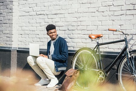 handsome african american freelancer working with laptop near bicycle at white brick wallの写真素材