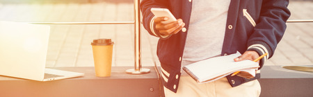 partial view of teleworker with documents, smartphone, laptop and coffee to go in city with sunlight