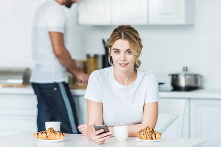 beautiful young woman using smartphone and smiling at camera while boyfriend preparing breakfast behindの写真素材