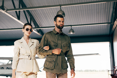low angle view of fashionable young couple in jackets walking near airplane in hangar