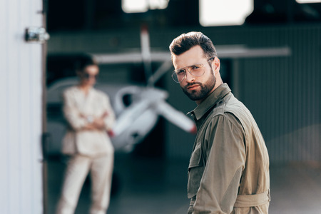 portrait of handsome man in eyeglasses and jacket looking at camera while his girlfriend standing in hangar with planeの写真素材