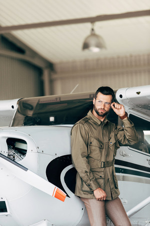 handsome young man in green jacket and eyeglasses looking away near airplaneの写真素材