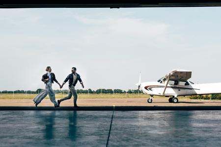distant view of stylish young couple in leather jackets holding hands and running near planeの写真素材
