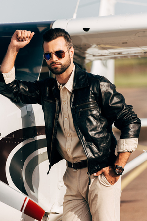 serious fashionable male pilot in leather jacket and sunglasses standing near airplaneの写真素材