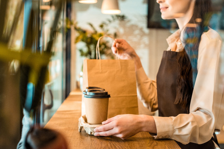 cropped image of waitress in apron holding disposable coffee cups and paper bag in cafeの写真素材