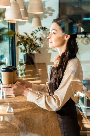 side view of beautiful waitress in apron holding disposable coffee cups in cafeの写真素材