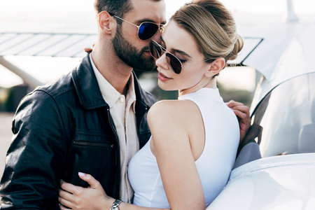 stylish man in leather jacket and sunglasses embracing attractive girlfriend near planeの写真素材