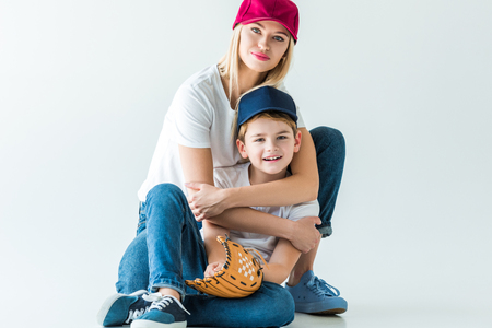 smiling mother hugging son and sitting on floor with baseball glove on whiteの写真素材