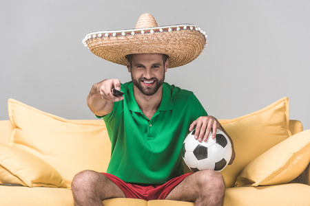 smiling man in mexican sombrero watching football match while sitting on yellow sofa with ball and remote control on greyの写真素材