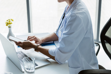 partial view of female doctor using smartphone at table with laptop in officeの写真素材