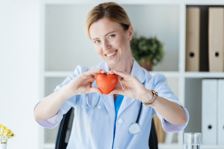 selective focus of female doctor in white coat showing heart symbol at table in officeの写真素材