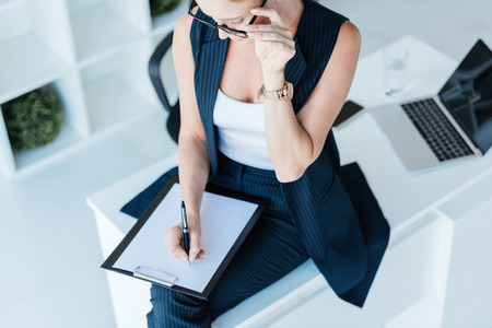 high angle view of businesswoman writing in clipboard on table with laptop in officeの写真素材