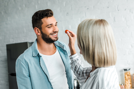 girlfriend feeding smiling handsome boyfriend with cherry tomato in kitchenの写真素材