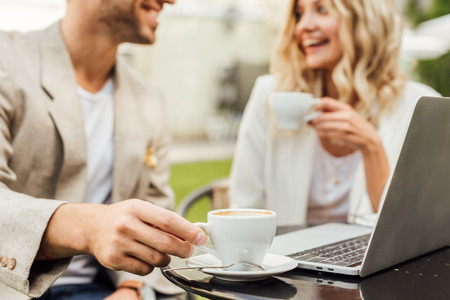 Cropped image of couple in autumn outfit sitting at table with laptop and drinking coffee in cafeの写真素材