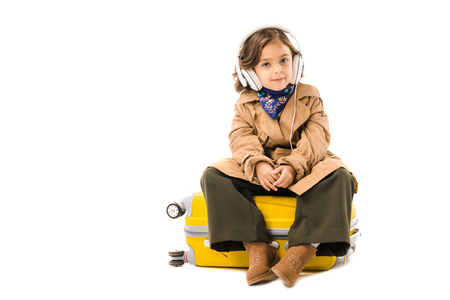 Adorable little child in trench coat listening music with headphones and sitting on yellow suitcase isolated on white backgroundの写真素材