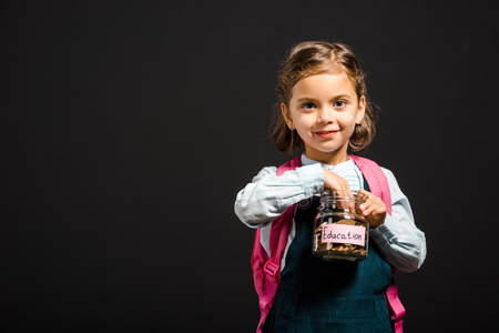 Schoolgirl with backpack holding glass jar with savings for education isolated on black backgroundの写真素材